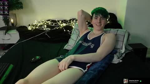 Media: Video of a young man with short green hair, fair skin, and a slim build, wearing a navy blue and white athletic tank top, reclining on a bed with black sheets, surrounded by fairy lights.