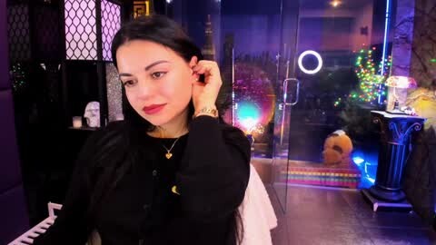 Media: Video of a stylish young woman with dark hair and fair skin, wearing a black top and red lipstick, sitting at a piano in a dimly lit room with neon lights, a ring light, and a plush toy.
