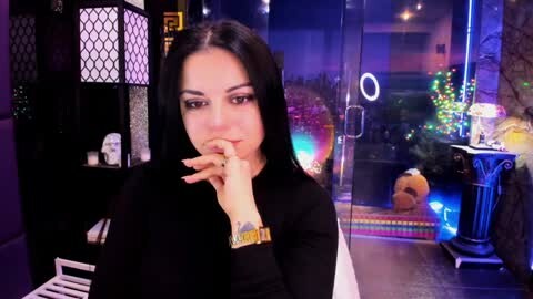 Video of a woman with long black hair, wearing a black top, smoking a cigarette, in a dimly-lit room with a glass wall, colorful lights, and a plush toy.