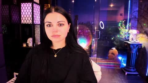 Media: Video of a woman with long black hair, fair skin, and a serene expression, wearing a black robe, standing in a dimly-lit, modern salon with glass doors and colorful lights in the background.