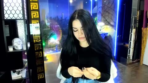 Media: Video of a young woman with long black hair, wearing a black top and blue jeans, looking at her phone, in a dimly lit room with a neon-lit cityscape background.