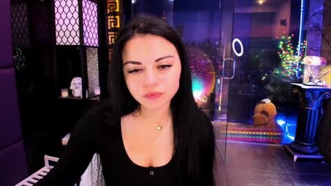 Media: Video of a young woman with long black hair, wearing a black top, sitting in a dimly lit room with a piano and colorful Christmas decorations in the background.