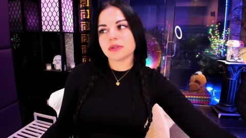 Media: Video of a woman with long black hair in a black top, sitting indoors by a window with vibrant neon lights and Christmas decorations in the background.