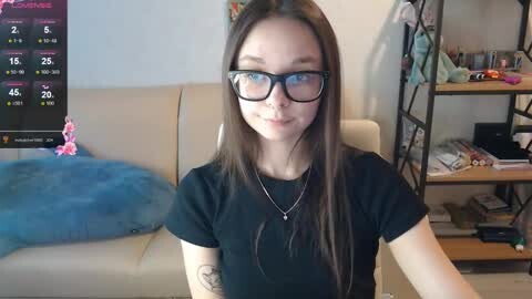 Media: A video of a young woman with long brown hair, wearing black-framed glasses and a black t-shirt, sitting in a cozy living room with a beige sofa, calendar, and bookshelf in the background.