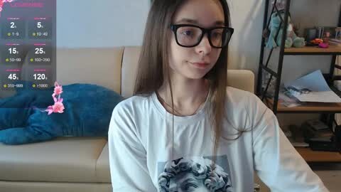 Media: Video of a young girl with long brown hair, glasses, wearing a white shirt with a graphic, sitting on a beige couch with a blue blanket and a pink plush toy.