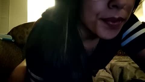 Media: A close-up video of a woman with light brown skin and dark hair, wearing a black and white striped shirt, licking her lips. Background features a brown teddy bear and a bed with beige sheets.