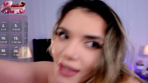 Media: Video of a young woman with light brown hair, light skin, and a slight smile, wearing a strapless top. Background shows a digital calendar and a blurred indoor setting.