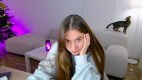 Media: Video of a young woman with long brown hair, resting her head on her hand, looking thoughtful in a modern living room with a Christmas tree, a grey sofa, and a lamp.
