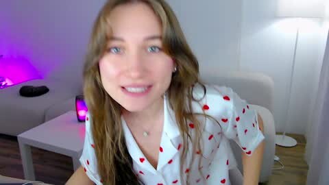 Media: Video of a young, smiling Caucasian woman with long, wavy brown hair, wearing a white shirt with red heart patterns, in a modern, brightly lit room with white furniture and a purple light.