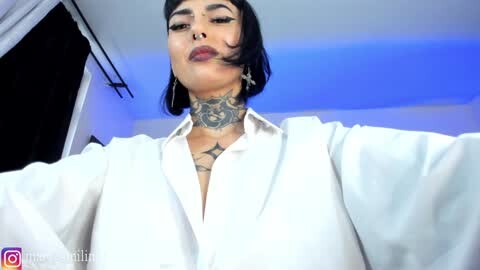 Media: Video of a woman with medium skin tone, short black hair, wearing a white blouse, tattoos on neck and chest, standing in a modern room with blue lighting.