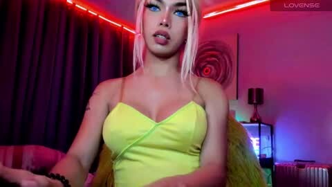 Video of a young woman with long, platinum blonde hair, wearing a tight, bright yellow spaghetti-strap dress, sitting in a dimly lit room with red and purple LED strip lights, visible tattoos on her arms.