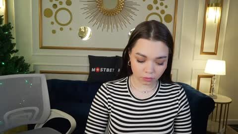 Video of a young woman with long brown hair in a black and white striped shirt, sitting in a modern living room with a blue sofa, Christmas tree, and golden wall decor.