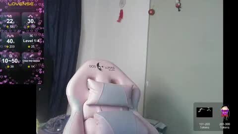 Media: A video captures a streamer's live stream on a pink gaming chair. The background shows a white wall with a red and gold tassel, a green plant, and a monitor displaying a game interface.