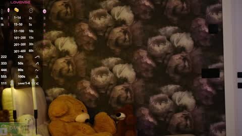 Media: Video of a teddy bear in a dark, textured wallpaper room with a digital screen displaying a camera feed. The bear is plush and brown, positioned on a soft surface.