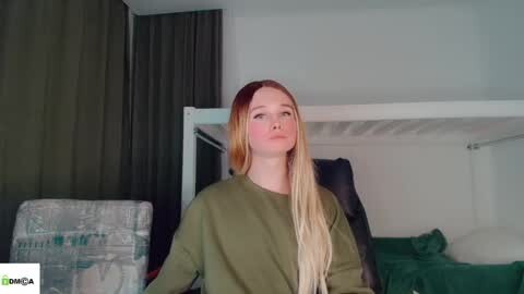 Media: Video of a fair-skinned, blonde-haired young woman in an olive green sweater, standing in a dimly lit bedroom with a white bunk bed and gray curtains.