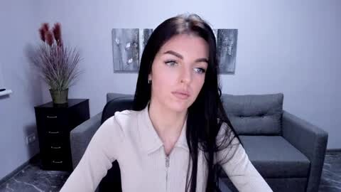 Media: Video of a fair-skinned woman with long black hair, wearing a cream-colored zip-up top, sitting on a grey couch. Minimalist living room background with a vase of tall red flowers on a black side table.