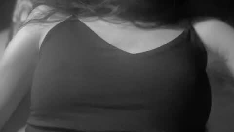 Media: Black and white video of a woman's upper torso, wearing a black sleeveless tank top, with wavy, shoulder-length hair partially visible. The image is slightly blurred, giving it a dreamy, vintage aesthetic.