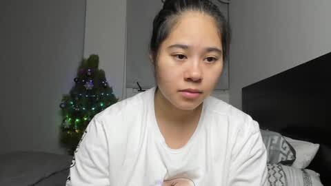 Media: Video of an Asian woman with long black hair in a ponytail, wearing a white t-shirt, sitting on a bed in a dimly lit room with a Christmas tree adorned with colorful lights in the background.