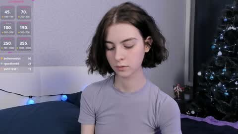 Media: A video of a young woman with short, dark brown hair and fair skin, wearing a light gray t-shirt, standing in a dimly lit room with a Christmas tree in the background.