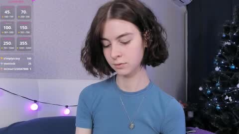 Media: Video of a young Caucasian woman with shoulder-length curly brown hair, wearing a light blue shirt, standing in a dimly lit room with Christmas decorations, including a string of purple lights and a decorated tree.