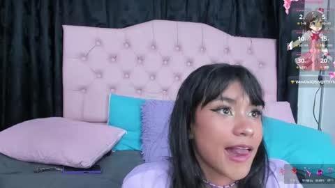 Media: Video of a Latina woman with long black hair, wearing glasses, lying on a bed with pink tufted headboard, surrounded by pastel pillows, and a TV showing a live stream.