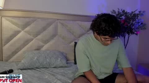 Media: A video of a young man with curly hair, wearing glasses and a light green t-shirt, sitting on a bed with a tufted headboard, in a dimly lit bedroom with a blue accent light.