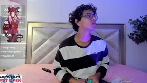 Media: Video of a young person with curly black hair, wearing glasses, a striped shirt, and a blue bracelet, sitting on a bed with a pink sheet, in a room with a white wall and a framed picture.