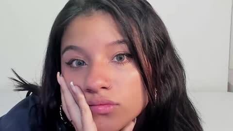 Media: Video of a young woman with medium brown skin, long dark hair, and expressive green eyes, resting her chin on her hand, wearing a dark top.
