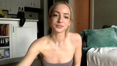 Media: Video of a young, fair-skinned woman with blonde hair, wearing a strapless top, sitting on a bed in a cozy, dimly lit room with white cabinets and green pillows.