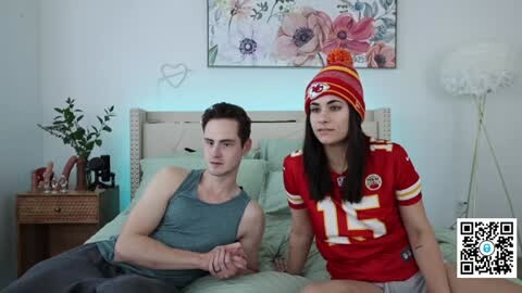 Media: Video of a man and woman in a bedroom; he's shirtless, muscular, and sits on a bed, while she, in a red football jersey and beanie, sits next to him.