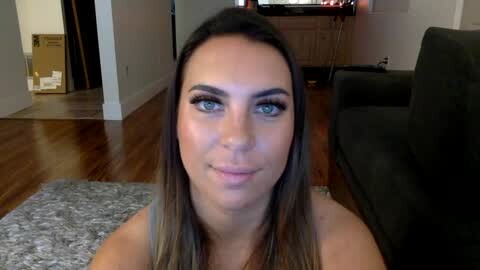 Media: Video of a smiling Caucasian woman with long brown hair and blue eyes, sitting on a gray rug in a living room with hardwood floors, dark furniture, and visible boxes.