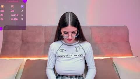 Media: Video of a young woman with long dark hair, wearing glasses and a white long-sleeved shirt with a logo, sitting on a brown couch against a pink wall.