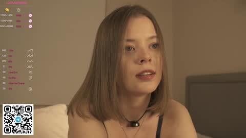 Media: Video of a young Caucasian woman with straight, shoulder-length brown hair, wearing a black top and a necklace. She's in a modern, beige-toned room with a TV screen displaying a QR code.