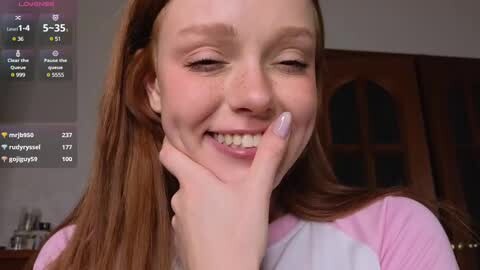 Media: A candid video of a young Caucasian girl with fair skin, long red hair, and a cheerful smile, holding her chin. She wears a pink and white top. Background includes wooden doors and a social media app screen.