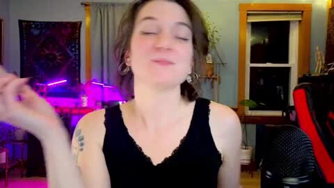 Media: A video of a young woman with fair skin, short brown hair, wearing a black tank top, playing with a cigarette in a dimly lit room with purple lights, plants, and a window.