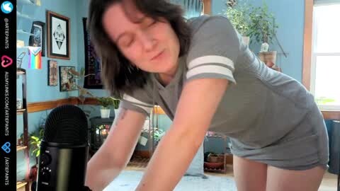 Media: Video of a middle-aged woman with short dark hair, wearing a grey t-shirt and short skirt, leaning over a black microphone in a cozy, sunlit room with framed artwork on blue walls.