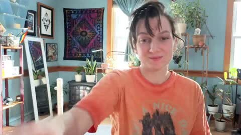 Media: Video of a young woman with fair skin and dark hair, wearing an orange t-shirt, sitting in a colorful, sunlit room with potted plants, wall art, and a wooden chair.