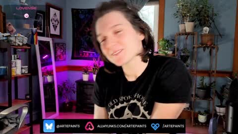 Media: Video of a smiling, fair-skinned woman with short, dark hair, wearing a black t-shirt, sitting in a dimly lit room with plants and framed art on the walls.