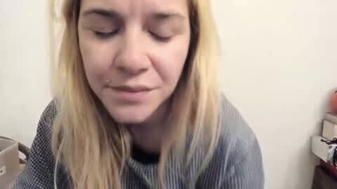 Media: Video of a young woman with light skin and long blonde hair, wearing a striped shirt, eyes closed, looking pensive, with a white wall and part of a desk in the background.