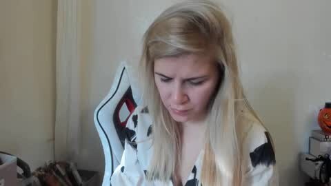 Media: A video of a young woman with long blonde hair, wearing a black and white patterned blouse, sitting at a desk in a dimly lit room.