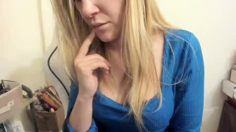 Media: Video of a young, light-skinned woman with long, straight blonde hair, wearing a blue ribbed top that reveals cleavage, contemplating with her hand on her chin. Background includes a beige wall, a shelf with books, and a white plastic bag.