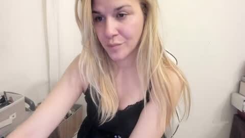 Media: Video of a fair-skinned, blonde woman in a black lace camisole, smiling slightly, leaning over a white counter in a kitchen.