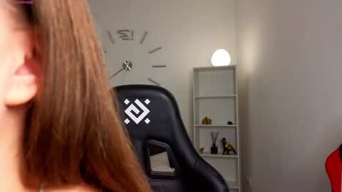 Media: Video of a woman with light skin and brown hair, wearing glasses, sitting in a black gaming chair. The background features a white wall with a modern, minimalist decor, including a spherical lamp and a bookshelf.