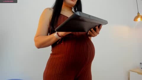 Media: Video of a pregnant woman in a sleeveless, ribbed, rust-colored dress holding a tablet, against a white wall with a modern pendant light.