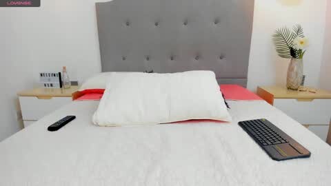 Media: A video of a minimalist bedroom with a white bed, grey upholstered headboard, white pillows, and a TV remote on the mattress.