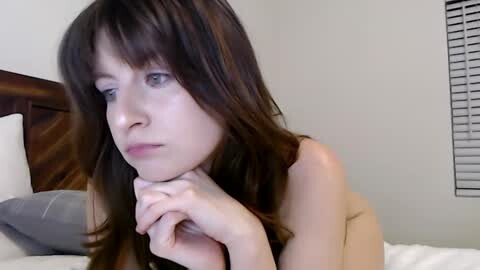 Media: Video of a nude young Caucasian woman with long brown hair, resting her chin on her hands, gazing introspectively. She is on a bed with white sheets and a wooden headboard, in a simple, sparsely decorated bedroom with a window with blinds.