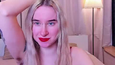 Media: Video of a smiling, fair-skinned, blonde-haired person with blue eyes, wearing red lipstick, and a tattoo on their left arm, standing in a dimly lit room with a white lamp and curtains.