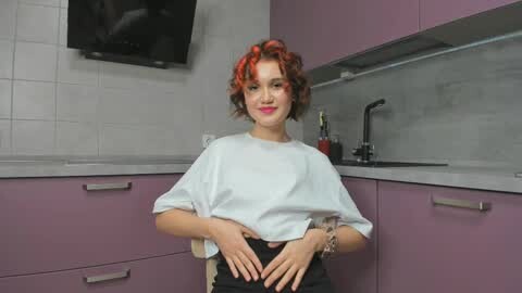 Media: Video of a young woman with curly red hair, wearing a white, loose-fitting top and black pants, standing in a modern kitchen with purple cabinets, white tiles, and a black TV.