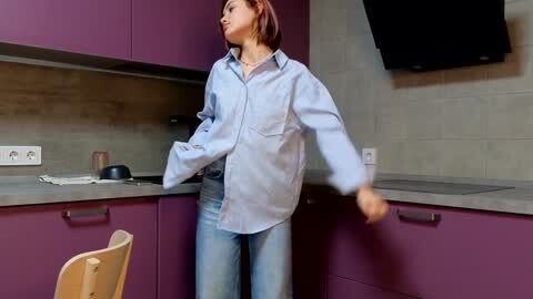 Media: Video of a young woman with shoulder-length auburn hair in a light blue button-up shirt and light-wash jeans, dancing in a modern kitchen with purple cabinets, grey backsplash, and a black wall-mounted oven.
