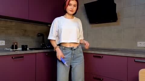 Media: A video of a young woman with short red hair, wearing a white crop top and high-waisted light blue jeans, standing in a modern kitchen with purple cabinets, a gray backsplash, and a black range hood.